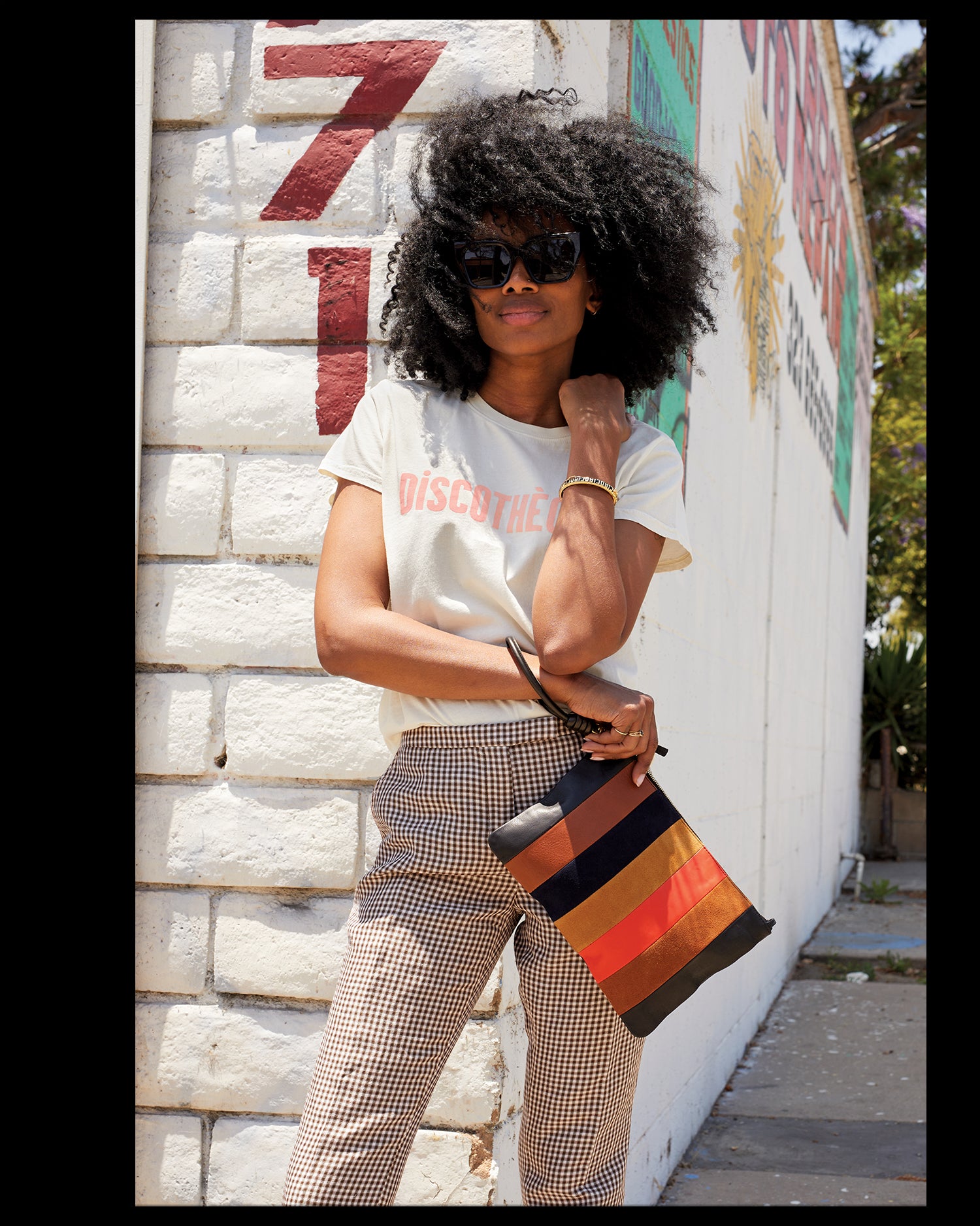 Mecca wearing our Discotheque tee and carrying the Suede / Nappa / Rustic Patchwork Flat Clutch with Tabs via a Black Cord Wristlet that's attached to the clutch.