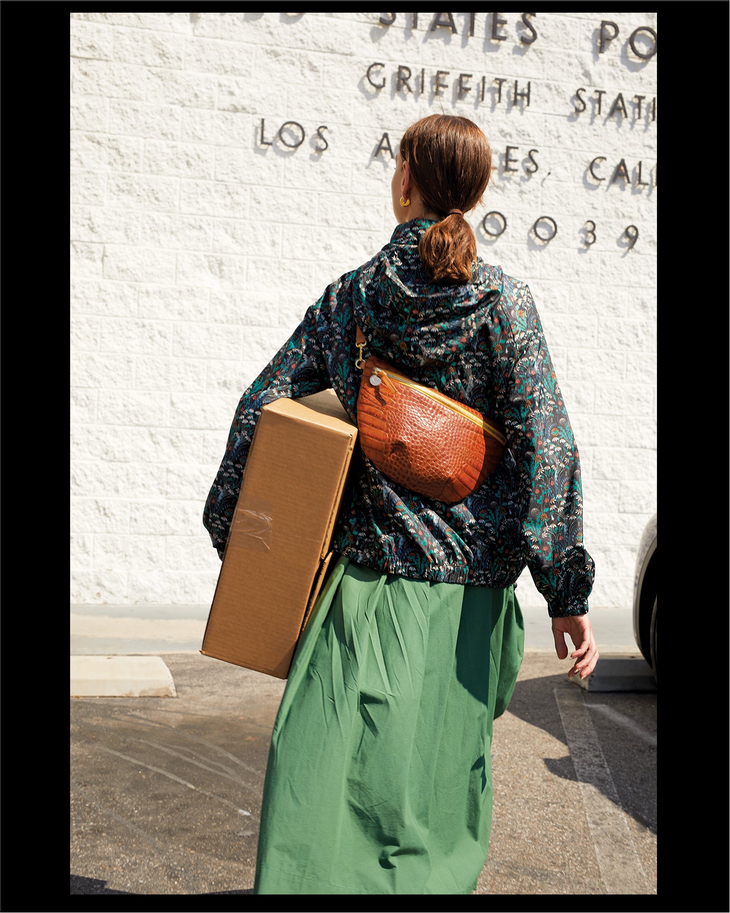 Frannie walking to the post office carrying a package and wearing the Cuoio Croco Grande Fanny Pack around her chest with the Grande Fanny pack hanging on the back of her body.
