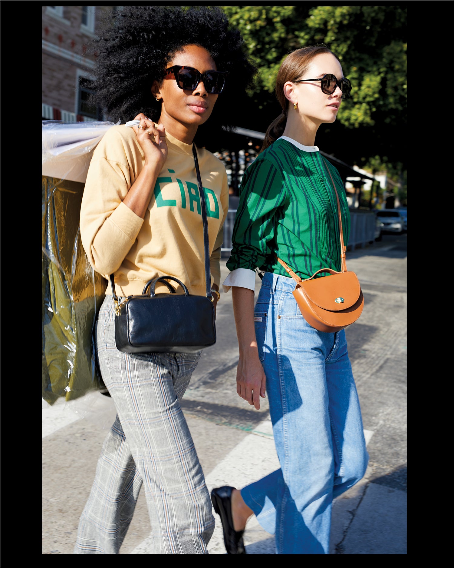 Mecca wearing the oat Ciao Le Drop Sweatshirt with plaid pants and Black Petit Claude across her chest. Model standing besides Mecca is wearing the Green & Black Stripe Le Tux with Jeans and Cuoio Elodie across her chest.