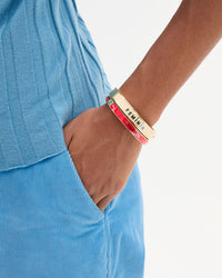 A close-up of a person wearing two stacked Le Bangle Bracelets. One with the word 'COMEDIE' on it, paired with a cream Le Bangle with the word 'FEMININ".