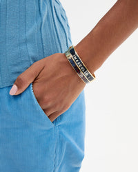 A close-up of a person's wrist stacking two Le Bangles that read "MASCULIN" and "TRAGEDIE".