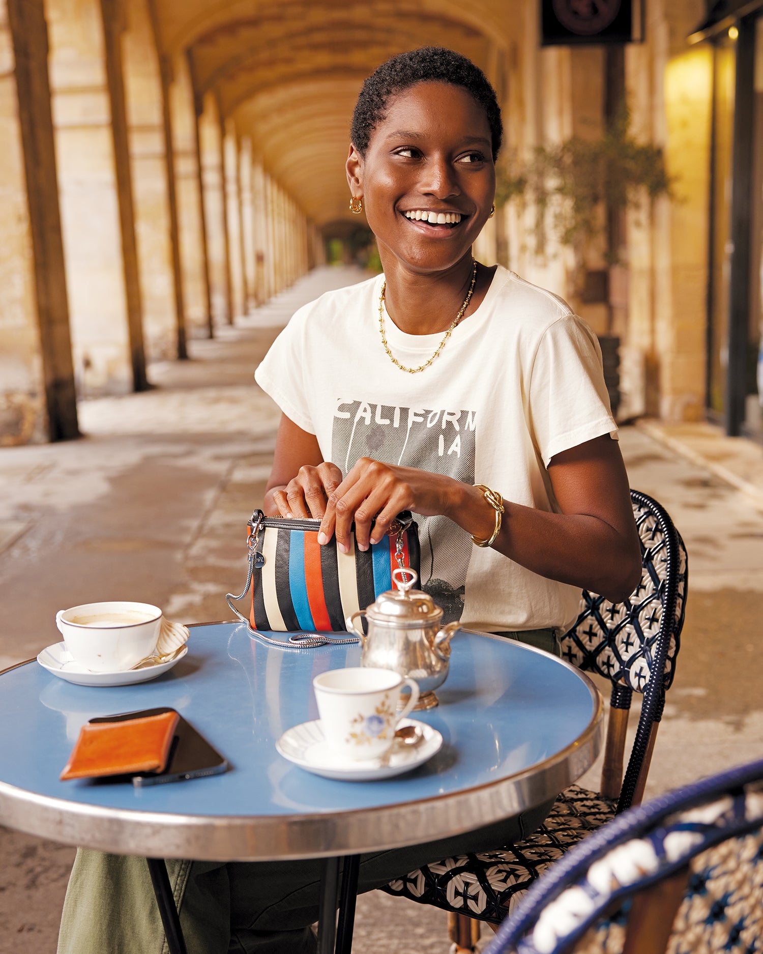 Model sitting in an outdoor French cafe holding the Wallet Clutch with Tabs in Striped Nappa with the SIlver Snake Chain Crossbody Strap attached to it.