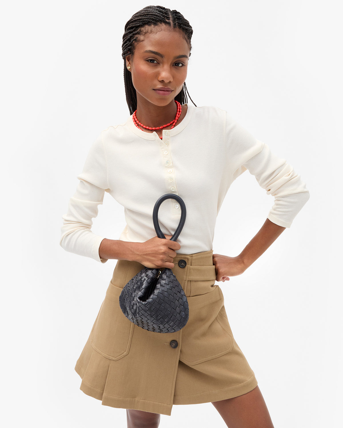 A model wearing a cream long-sleeve henley top, a beige wrap-style pleated skirt, and holding the Twilight Diagonal Woven Petit Moyen scrunched up by the tubular shoulder strap.
