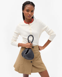 A model wearing a cream long-sleeve henley top, a beige wrap-style pleated skirt, and holding the Twilight Diagonal Woven Petit Moyen scrunched up by the tubular shoulder strap.