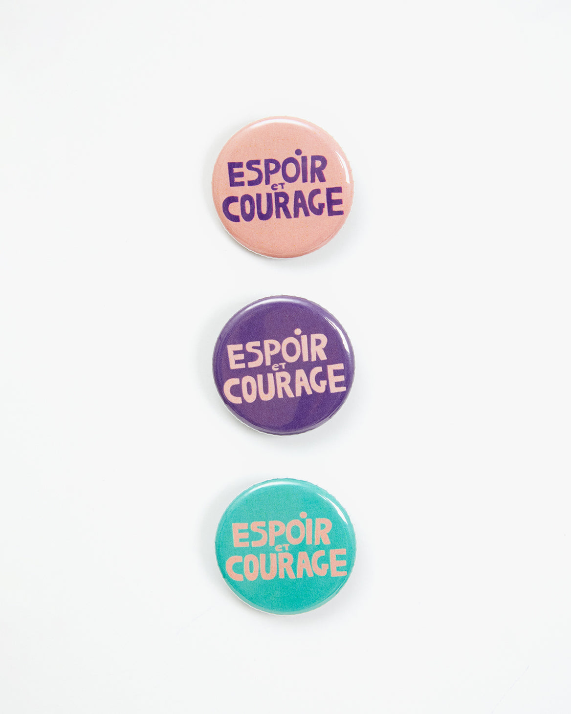 All Three Pins From Our CV x Giffords Canvas Totes With Pins. There are Three Pins with the words Espoir Et Courage on them, they come in colors light pink with purple writing, purple with light pink writing, and turquoise with light pink writing.