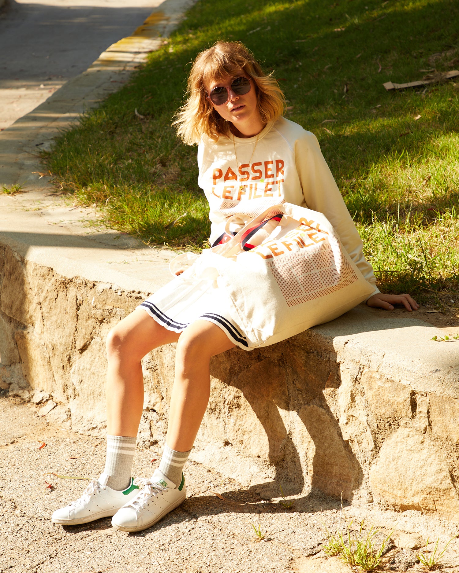 Lou wearing our CV x Racquet collab PASSER LE FiLET Sweatshirtand matching Canvas Jumbo Tote. Lou is wearing the sweatshirt with a cream tennis skirt and white Adidas tennis shoes and is sitting outside.