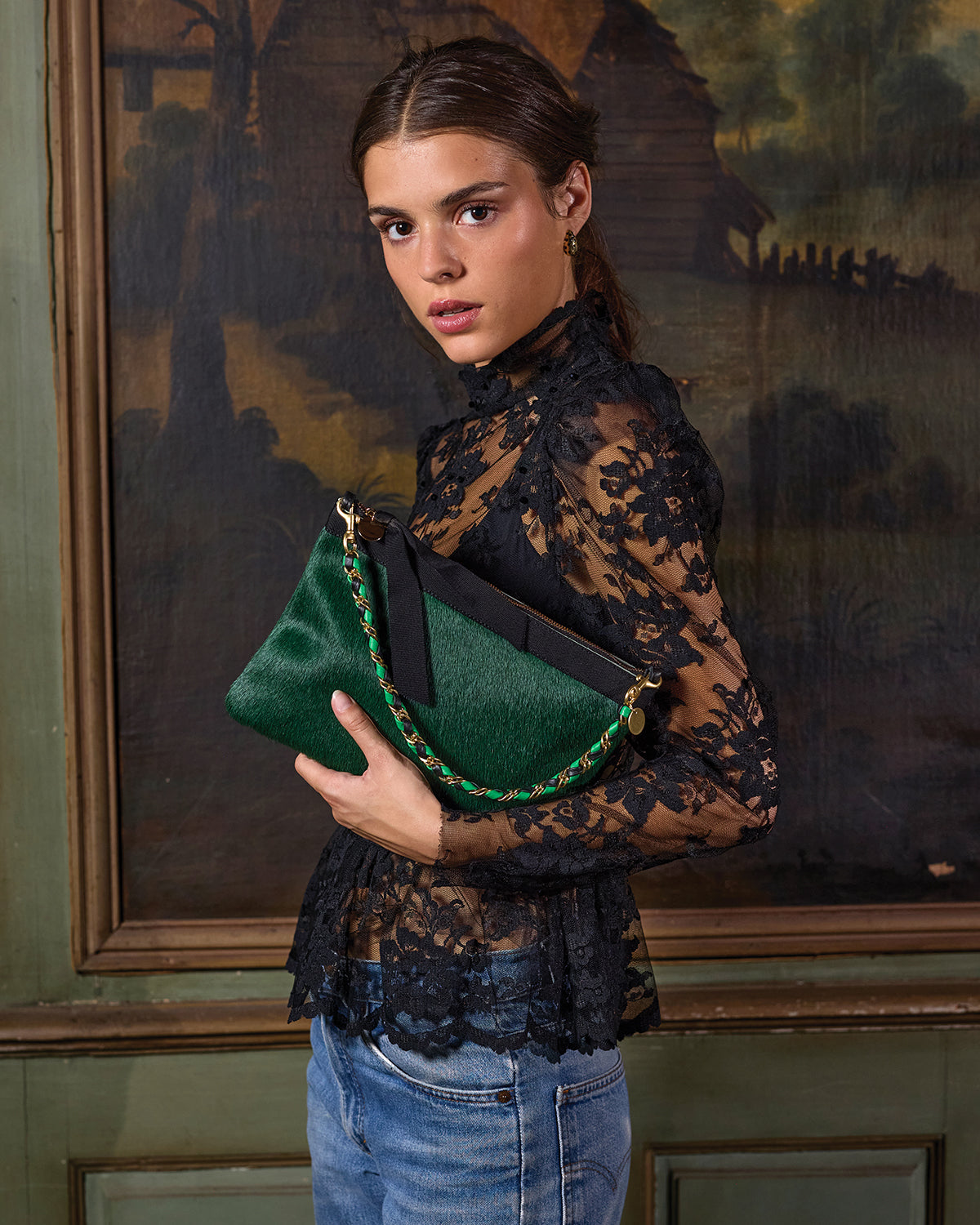 Model wearing a black lace top with blue jeans and holding the Bottle Green Hair-On Flat Clutch with Tabs with Black/Emerald Leather Wrapped Chain
 Shortie Strap attached to the clutch.  