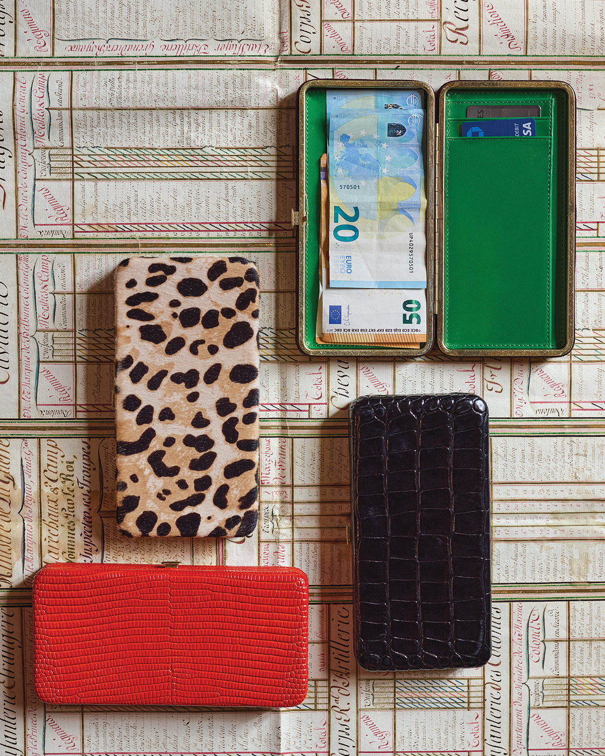 Le Tabacs in Red Embossed Lizard, Snow Leopard, Black Croco, and Truffle Suede which is open and is showing its green interior with foreign money inside. 