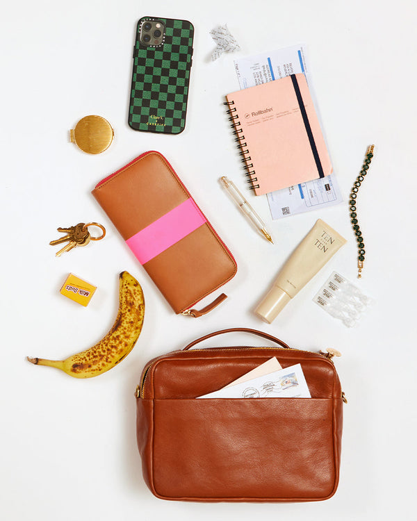Miel Mirabel laying flay on a white surface with an array of items that fit inside of it. These items include an iPhone, a small compact, a set of keys, a mini package of Milk Duds, a Banana, a full Zip wallet, a pen, a bracelet, a small notebook, a small lotion and a few letters.