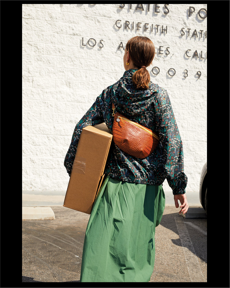 Frannie walking to the post office carrying a package and wearing the Cuoio Croco Grande Fanny Pack around her chest with the Grande Fanny pack hanging on the back of her body.