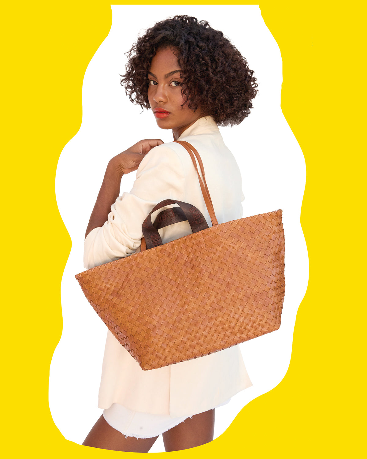 Model wearing a cream blazer and carrying the Natural Woven Diagonal Bateau Tote over her shoulder. 