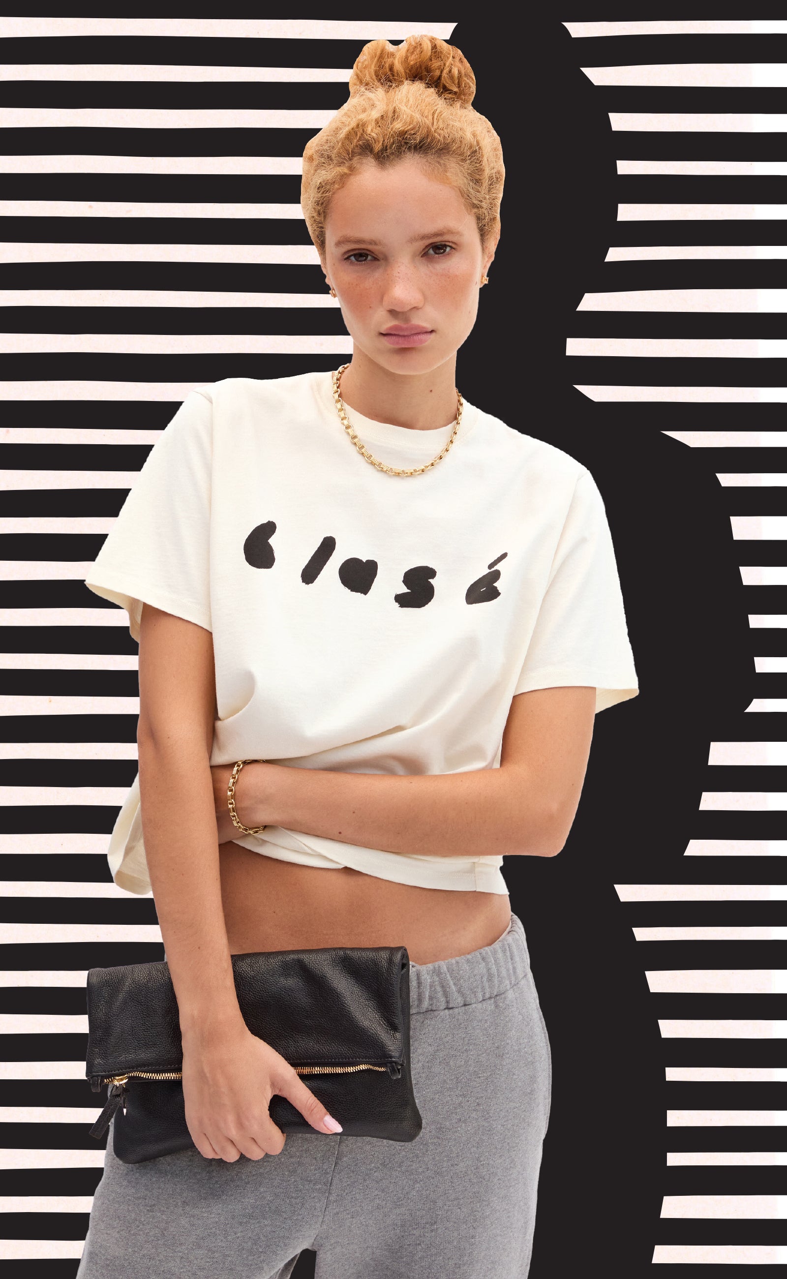 Model wearing the Cream Blasé Original Tee and holding the Black Petit Caviar Foldover Clutch with Tabs. 