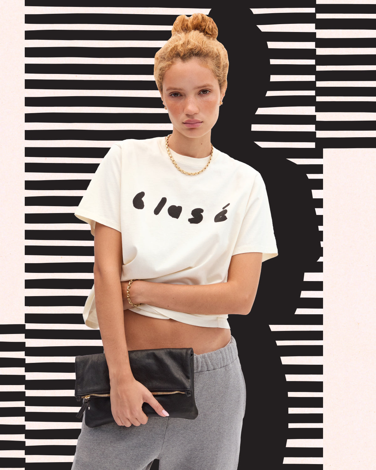 Model wearing the Cream Blasé Original Tee and holding the Black Petit Caviar Foldover Clutch with Tabs. 