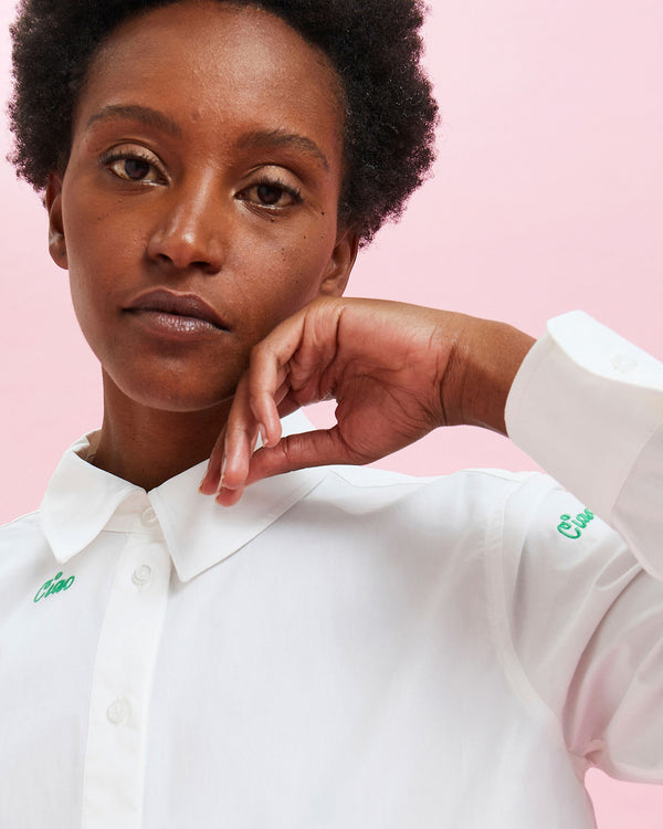 model with her hand to her face wearing the White w/ Green Embroidered Ciao Button Up Shirt buttoned all the way up 