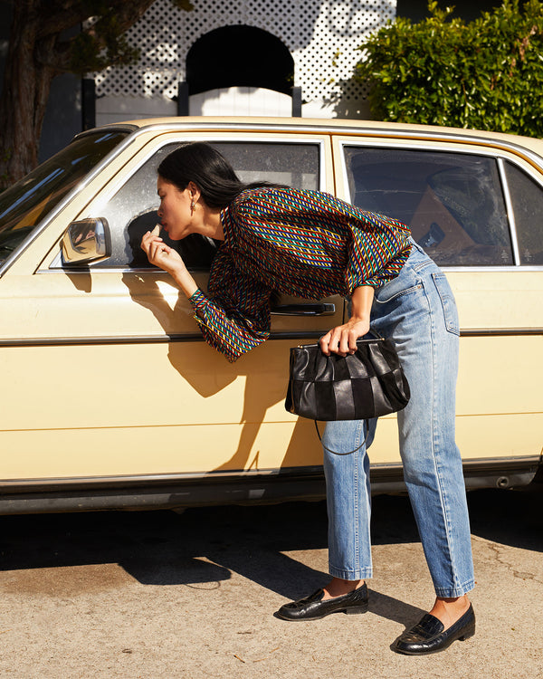Sandra touching up her lipstick in the side mirror of a car while holding on to the Black Suede & Nappa Belle