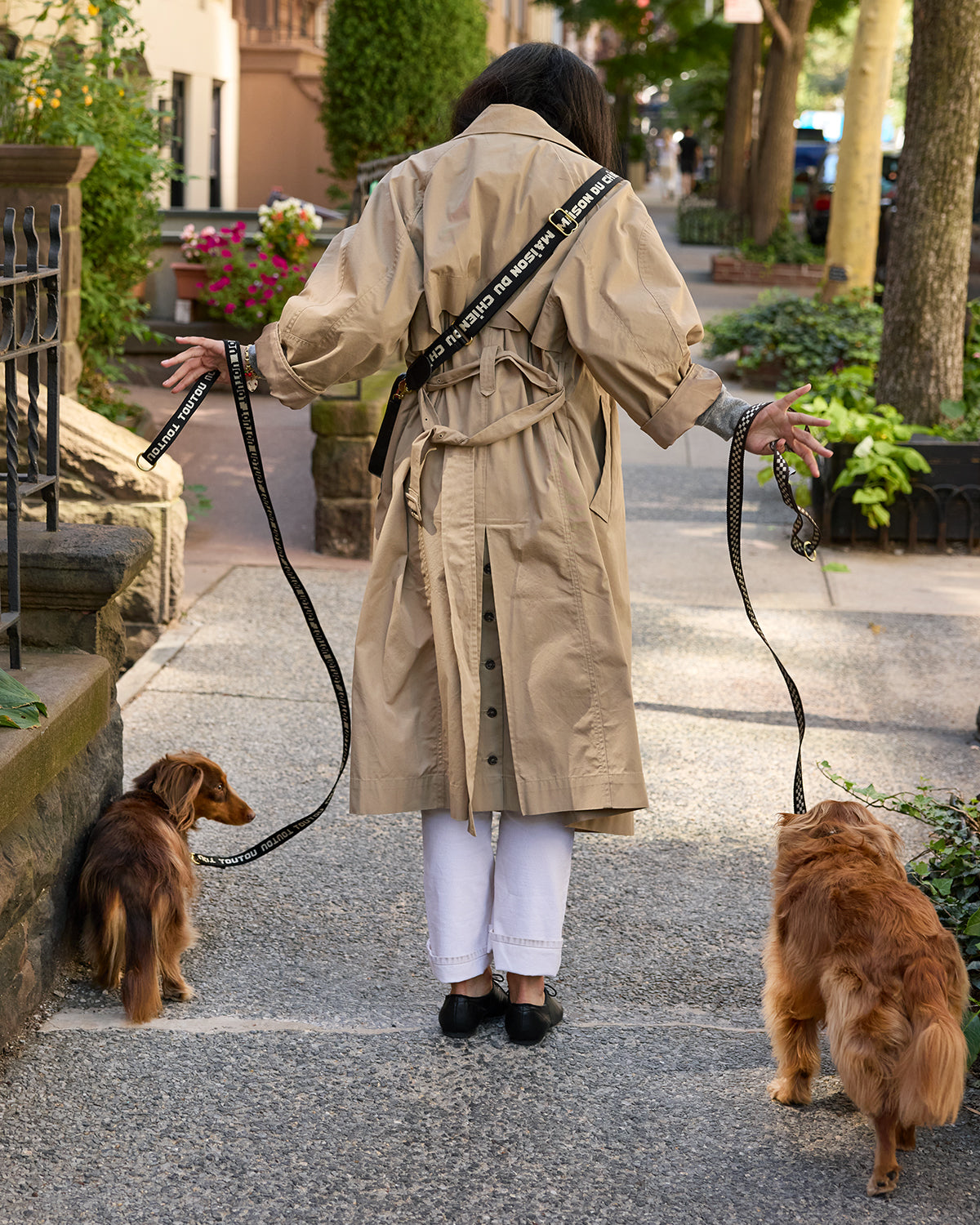Back of a model wearing a trench coat and walking two dogs with the CV x The Dogist leashes. You can see she's carrying the CV x The Dogist Poche Sportif crossbody bag as the strap is showing in the back. 