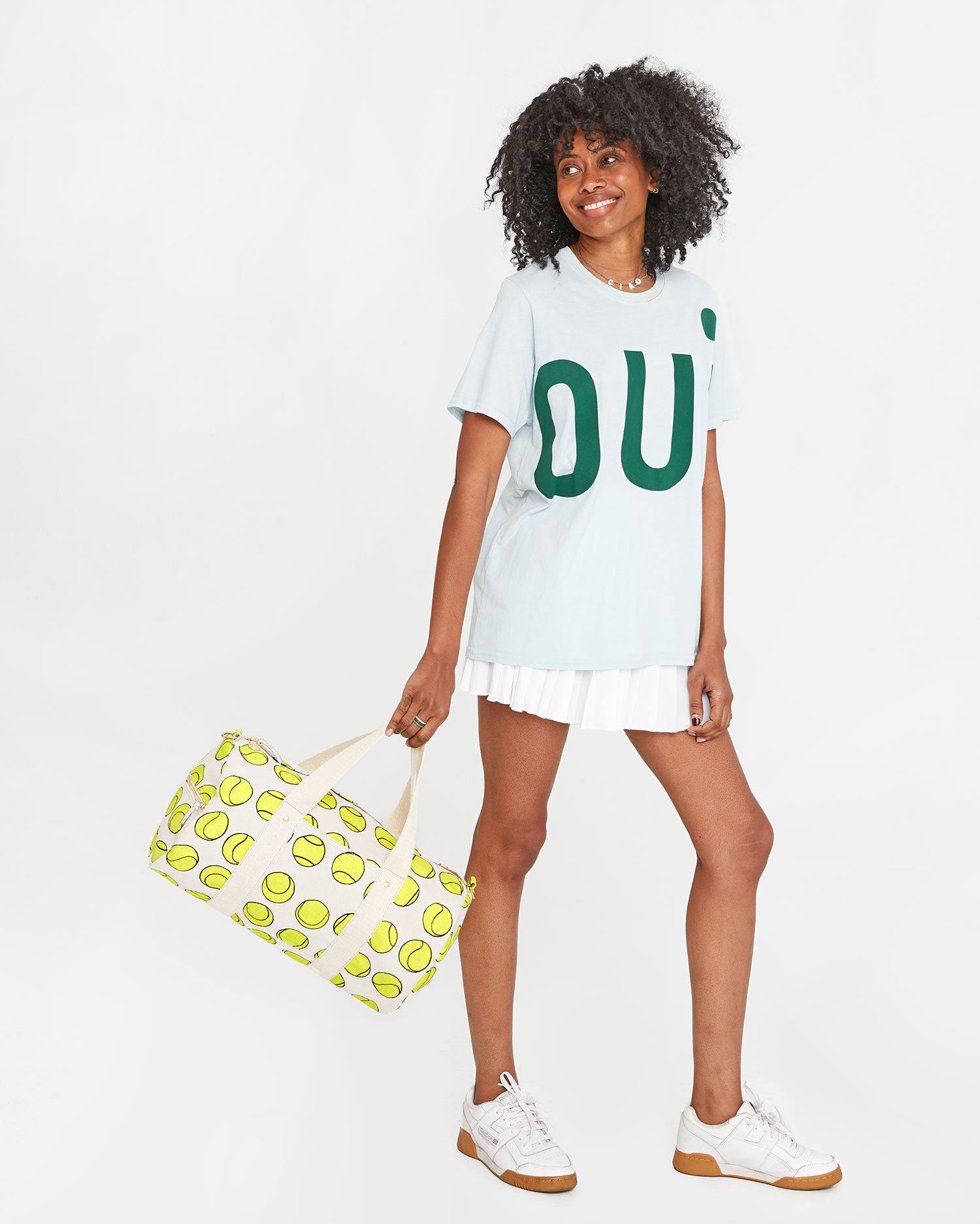 Mecca wearing a white tennis skirt with the sky blue grand oui t shirt . she's carrying the Natural w/ Tennis Balls Duffle