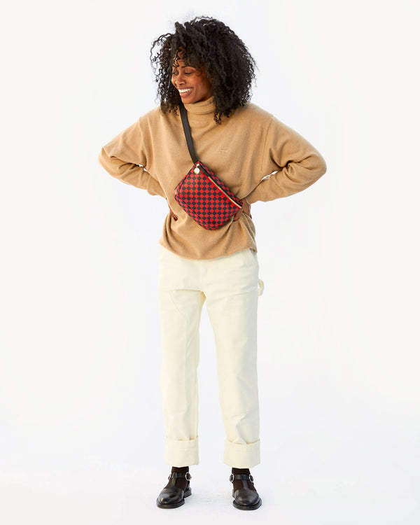 Mecca wearing the Cherry Red & Navy Checkers Fanny Pack across her chest. She has on white pants and a tan sweater