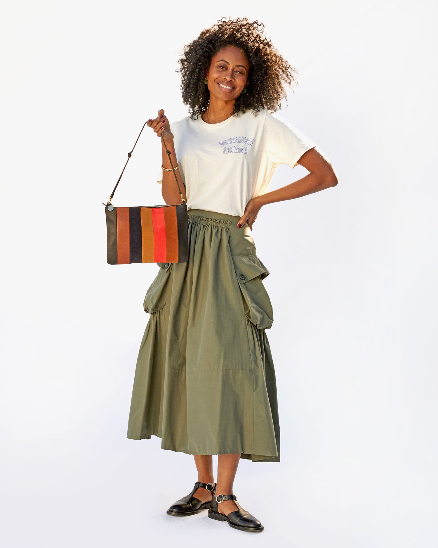 Mecca in a tshirt and an army green skirt holding a flat clutch with tabs by the Black Thin Knotted Shoulder Strap