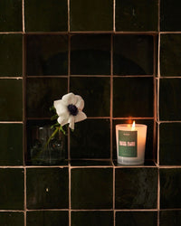 a lit Roen Candle in the scent Hotel Flori next to a single flower in a glass on  a tiled background
