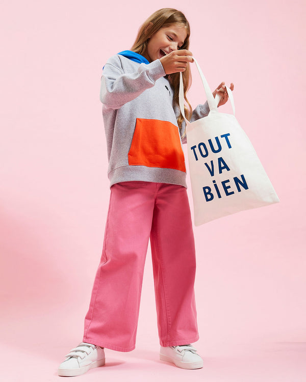 model wearing the Grey w/ Poppy & Cobalt Kids Hoodie with pink pants and white sneakers, holding the kids tote bag