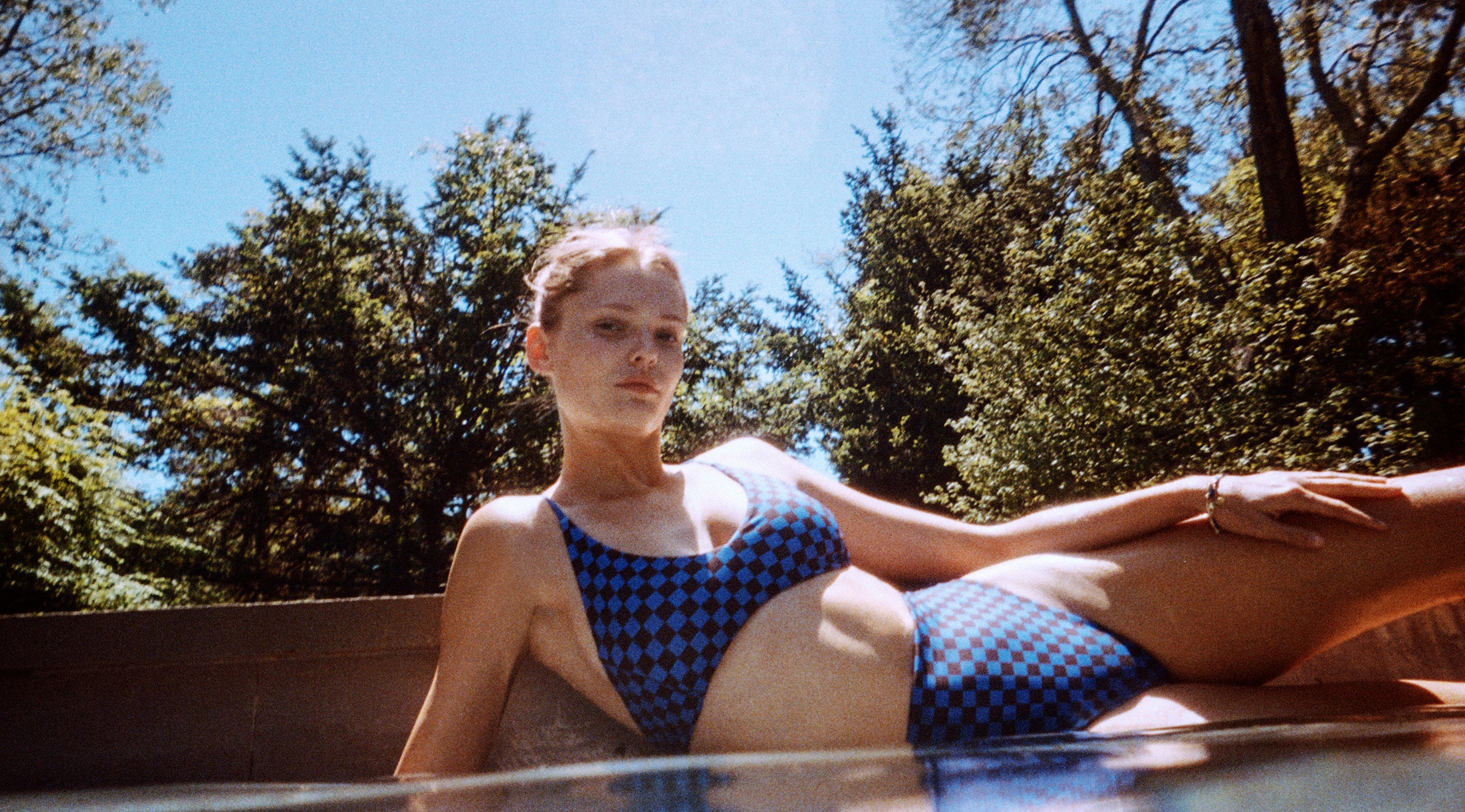 Model laying by the pool wearing a 2-Piece CV x Left On Friday swimsuit in our checkered print. 