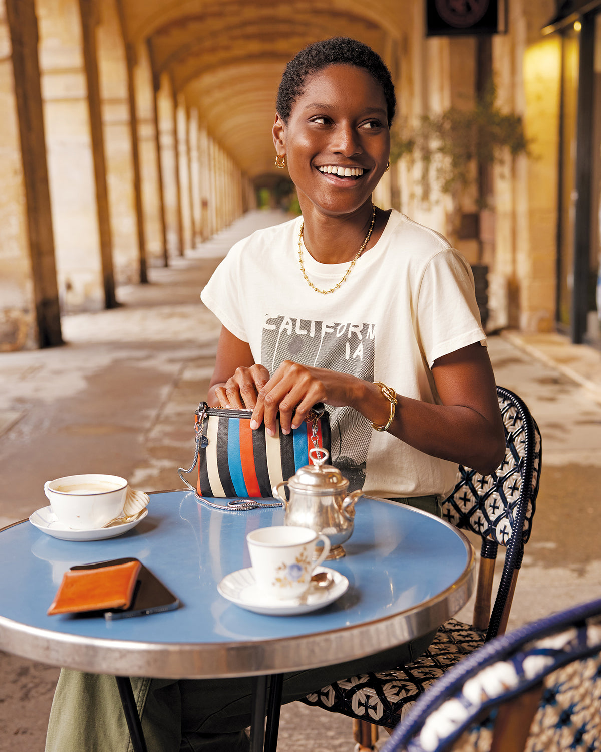 Model sitting in an outdoor French cafe holding the Wallet Clutch with Tabs in Striped Nappa with the SIlver Snake Chain Crossbody Strap attached to it. 