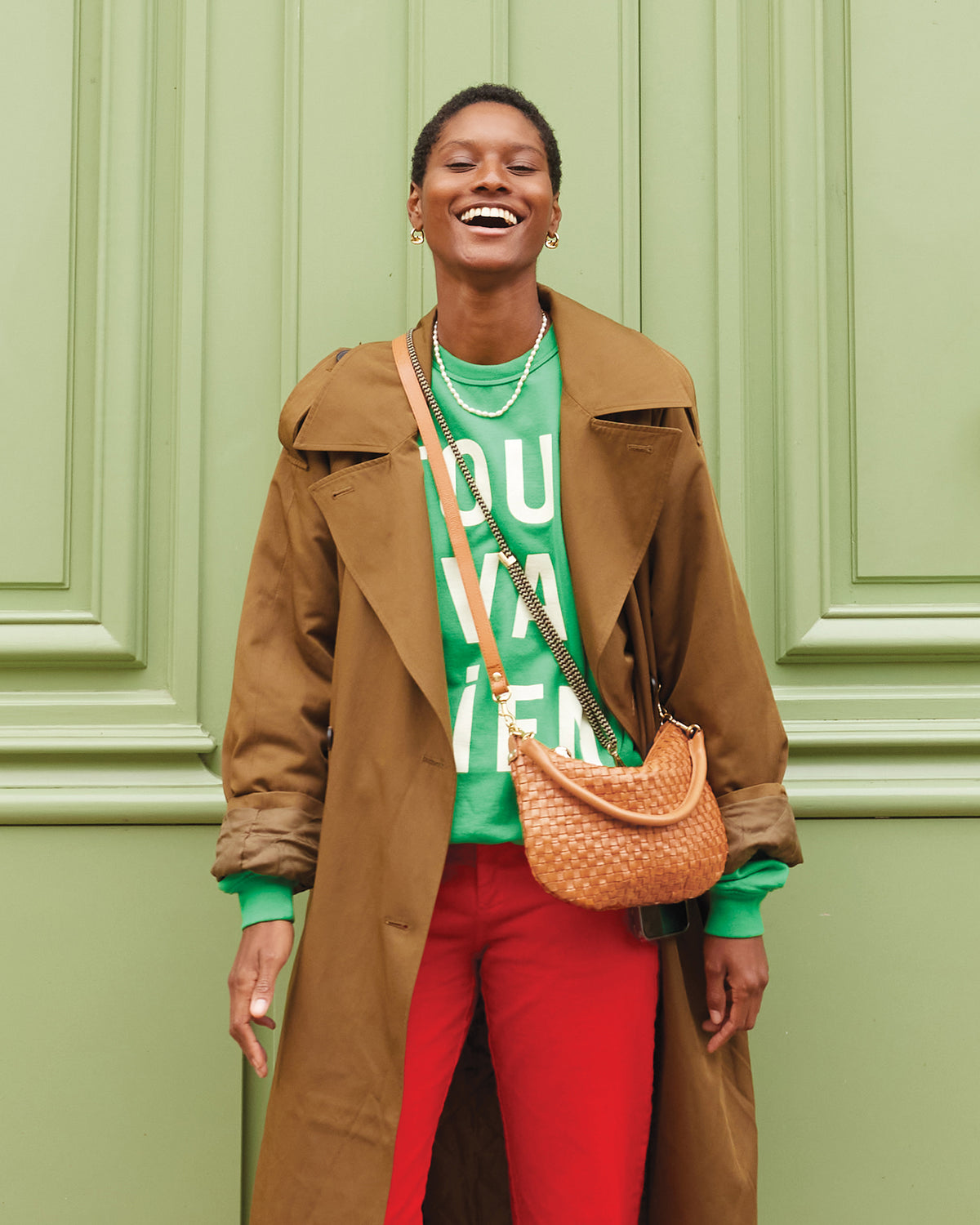 Model wearing the Green Tout Va Bien Oversized Sweatshirt under a trench coat and carrying the Natural Woven Petit Moyen as a crossbody bag. 