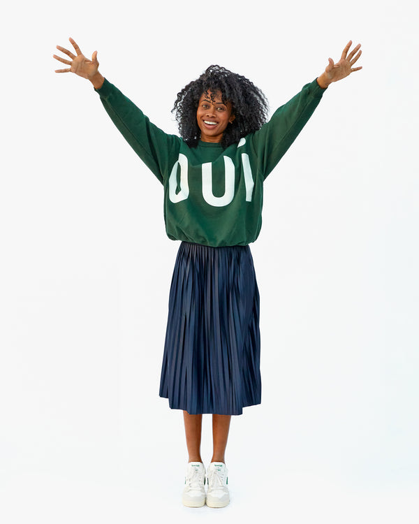 Mecca in a blue pleated skirt, the Forest Grand Oui Oversized Sweatshirt and white sneakers with her arms raised above her head