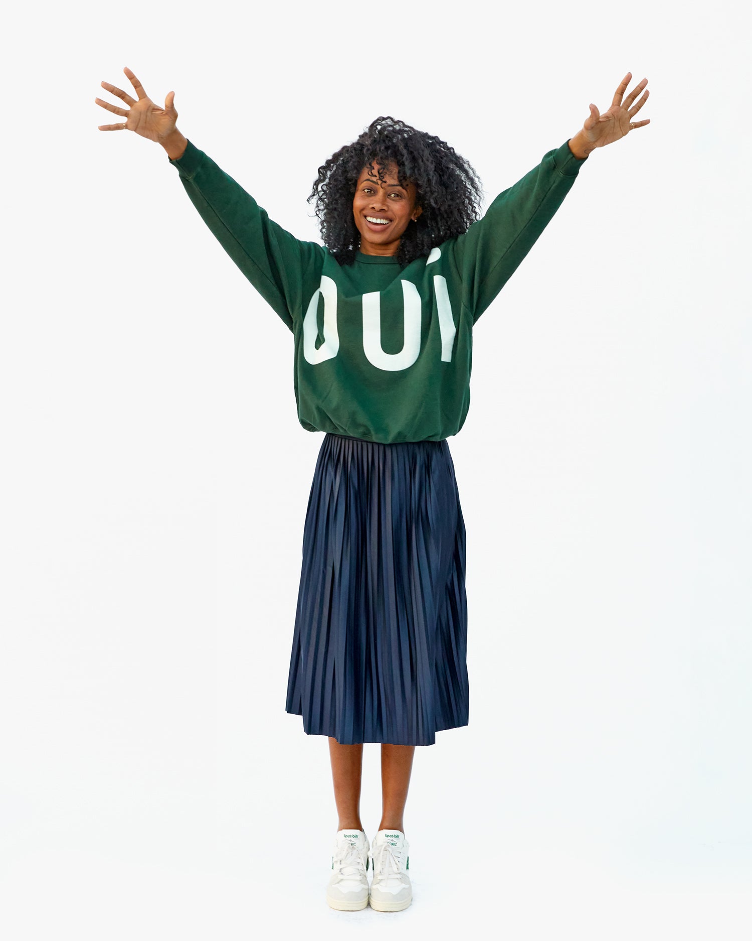 Mecca in a blue pleated skirt, the Forest Grand Oui Oversized Sweatshirt and white sneakers with her arms raised above her head