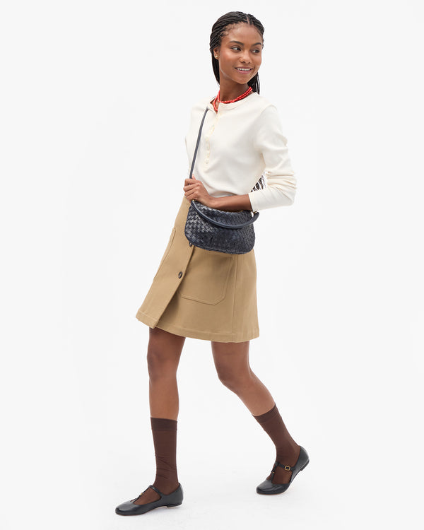 A model wearing a  white long-sleeve top, a beige wrap-style skirt, dark brown socks, and black flats, carrying the Twilight Diagonal Woven Petit Moyen as a crossbody bag. 