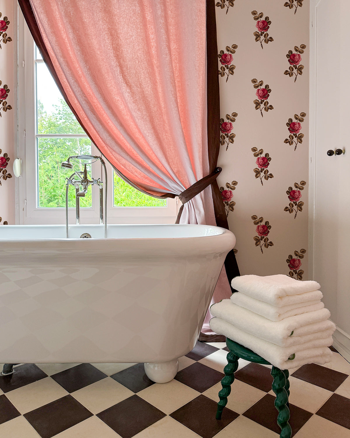 Bathroom with a white tub, green stool with towels, pink with chocolate border curtain, and 'Corsage' Wallpaper in Petal Brown.