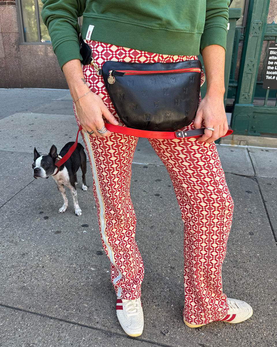 Person wearing the Clare V. x The Dogist Black Deboss Grande Fanny w/ Adjustable Webbing Strap  around their waist with their Bosten Terrier in the back wearing a CV x The Dogist collar and leash. 