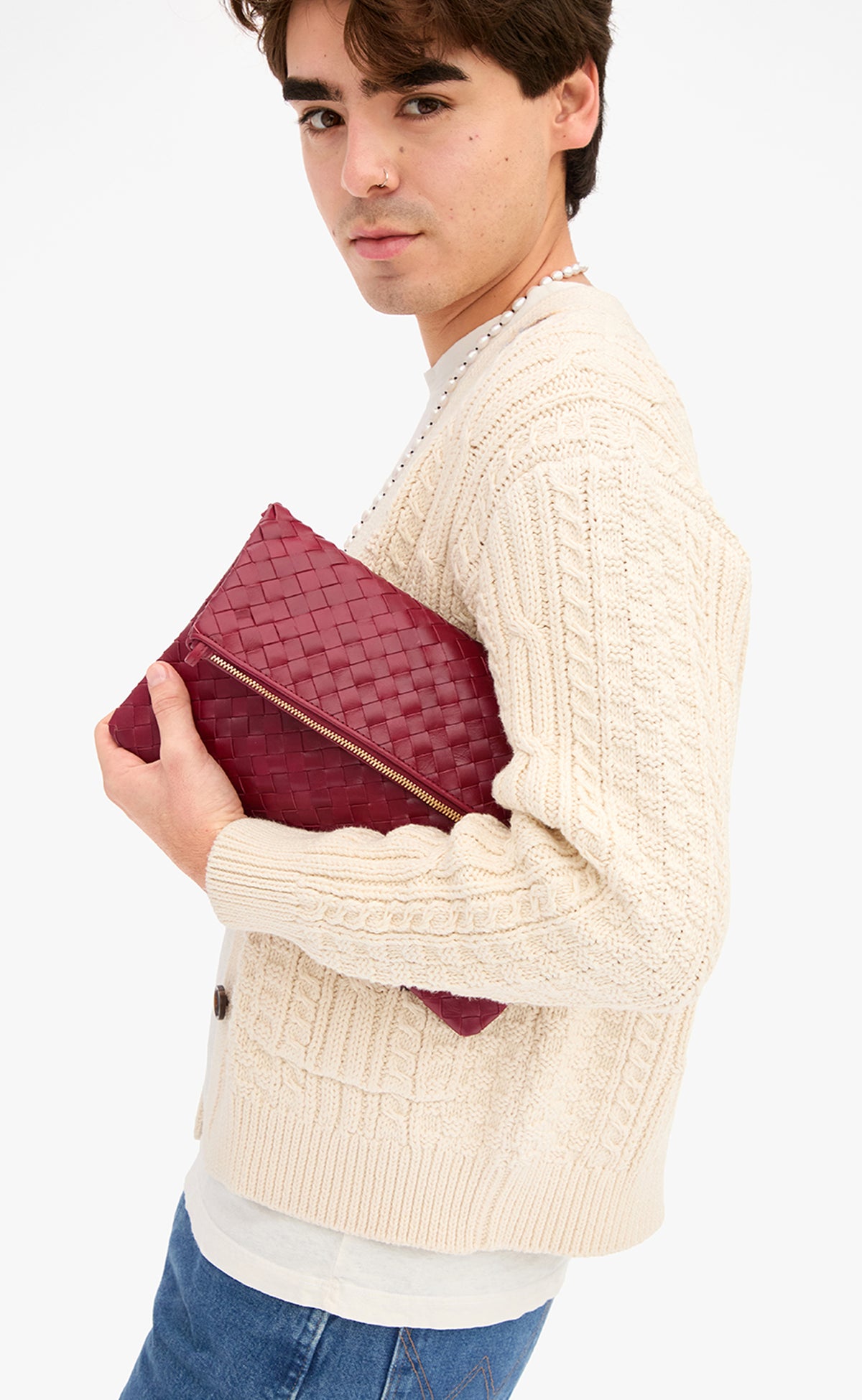 Male model wearing a cream cable knit cardigan with blue jeans and holding the Garnet Diagonal Woven Foldover Clutch w/ Strap as a foldover clutch without the strap. 
