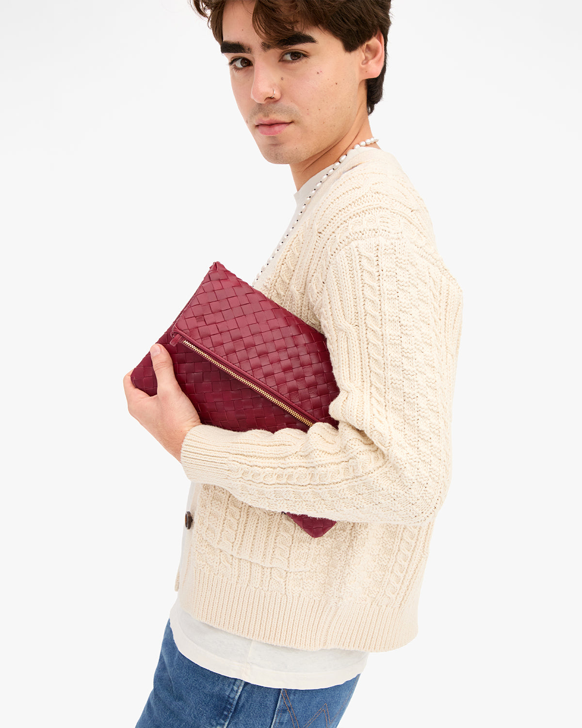 Male model wearing a cream cable knit cardigan with blue jeans and holding the Garnet Diagonal Woven Foldover Clutch w/ Strap as a foldover clutch without the strap. 
