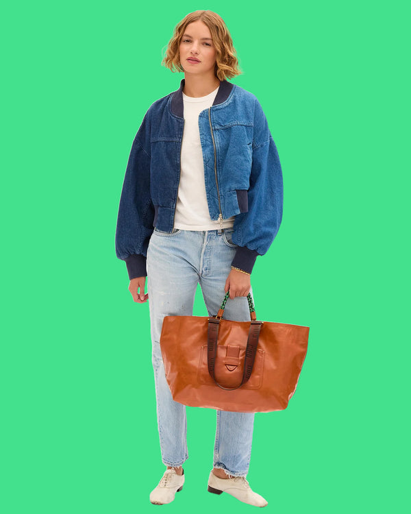 Model wearing jeans, a white t-shirt, and jean bomber jacket carrying the Camel Grande Bateau Tote by the top handle. 