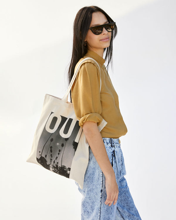 Aurelia is wearing the Natural Oui Canvas Store Tote on her shoulder with acid wash denim pants and a mustardy tan shirt
