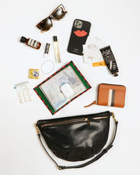 Black Grande Fanny showing the items that it can hold. These items include sunglasses, an aesop sample, chapstick, perfume, gum, an iphone, hand cream, a passport sleeve, airpods and a petit zip wallet