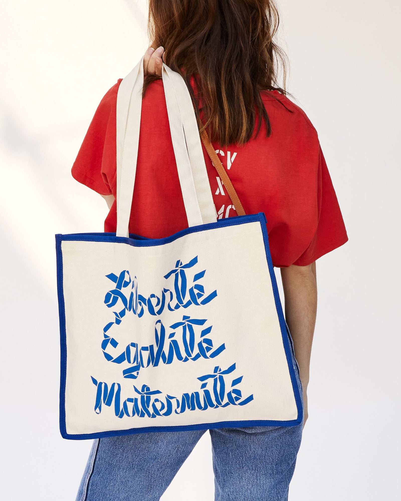 Back of Our CV x EMC Liberté, Egalité, Maternité Heavy Tee in Poppy on Frannie As She's Holding Our Liberte, Egalite, Maternite Cream with Blue Tote Over Her Shoulder