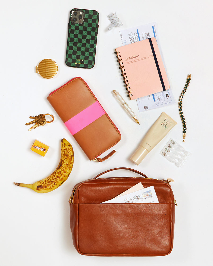 Miel Mirabel laying flay on a white surface with an array of items that fit inside of it. These items include an iPhone, a small compact, a set of keys, a mini package of Milk Duds, a Banana, a full Zip wallet, a pen, a bracelet, a small notebook, a small lotion and a few letters. 