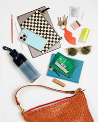 The natural woven moyen messenger with an array of items that fit inside of it. These items include a small water bottle, two pencils, an ipad, a wallet clutch with tabs, an iphone, a set of keys with a lips key fob, a small set of matches, a stick sunscreen, sunnies, a small notebook, a coin clutch, a back of gum and a lipstick.